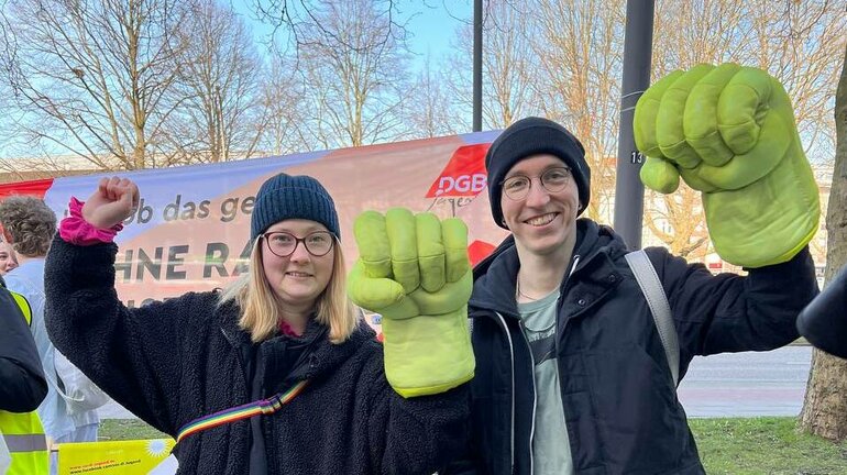 Zwei Personen blicken mit erhobenem linken Arm über dem jeweils ein Arbeiterfaust Handschuh steckt in die Kamera beim verdi Jugendstreiktag der Tarifrunde öffentlicher Dienst Bund und Kommunen 2023 in Hamburg