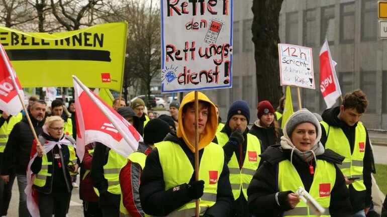 Am verdi Jugendstreiktag der Tarifrunde öffentlicher Dienst Bund und Kommunen 2023 in Kiel ziehen Demonstrierende mit gelben verdi Westen, Schildern und Fahnen durch die Straße. Eine Person trägt ein Schild auf dem steht: Rettet den Rettungsdienst