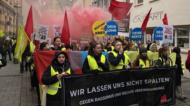 eine Gruppe Streikender trägt ein Banner mit der Aufschrift Wir lassen uns nicht spalten. In der Luft ist roter Rauch.