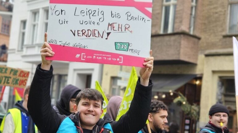 Ein streikender junger Mann trägt ein Schild, auf dem steht "Von Leipzig bis Berlin, wir wollen mehr verdien".