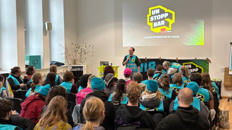 Eine große Gruppe Jugendlicher sitzt in einem Raum und hört einer präsentierenden Person zum Jugenstreiktag in Leipzig zu.