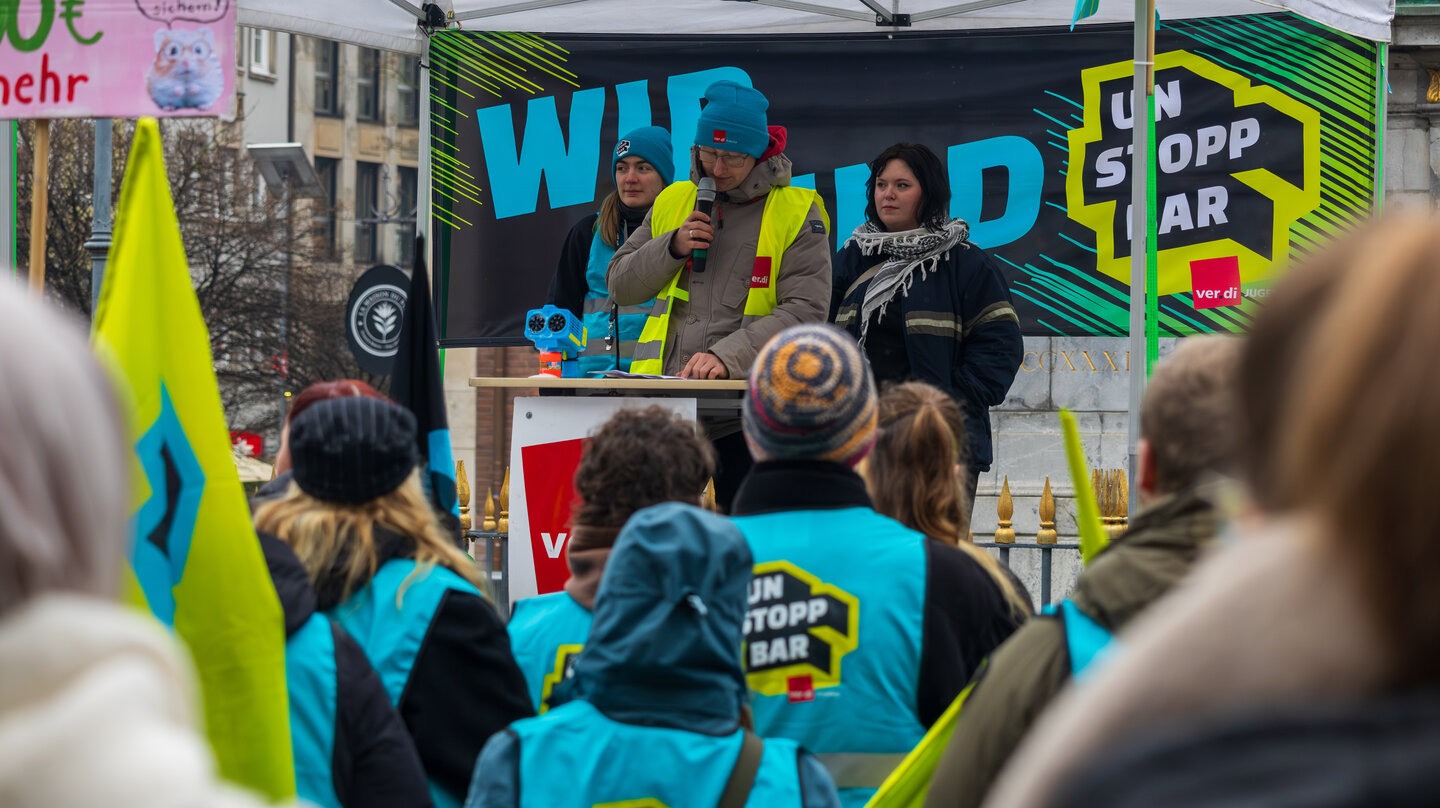 Auf einer Bühne steht eine Person in einer Streikweste vor einem Banner mit der Aufschrift "Wir sind Unstoppbar" und spricht zu einer Gruppe Personen in Streikwesten.